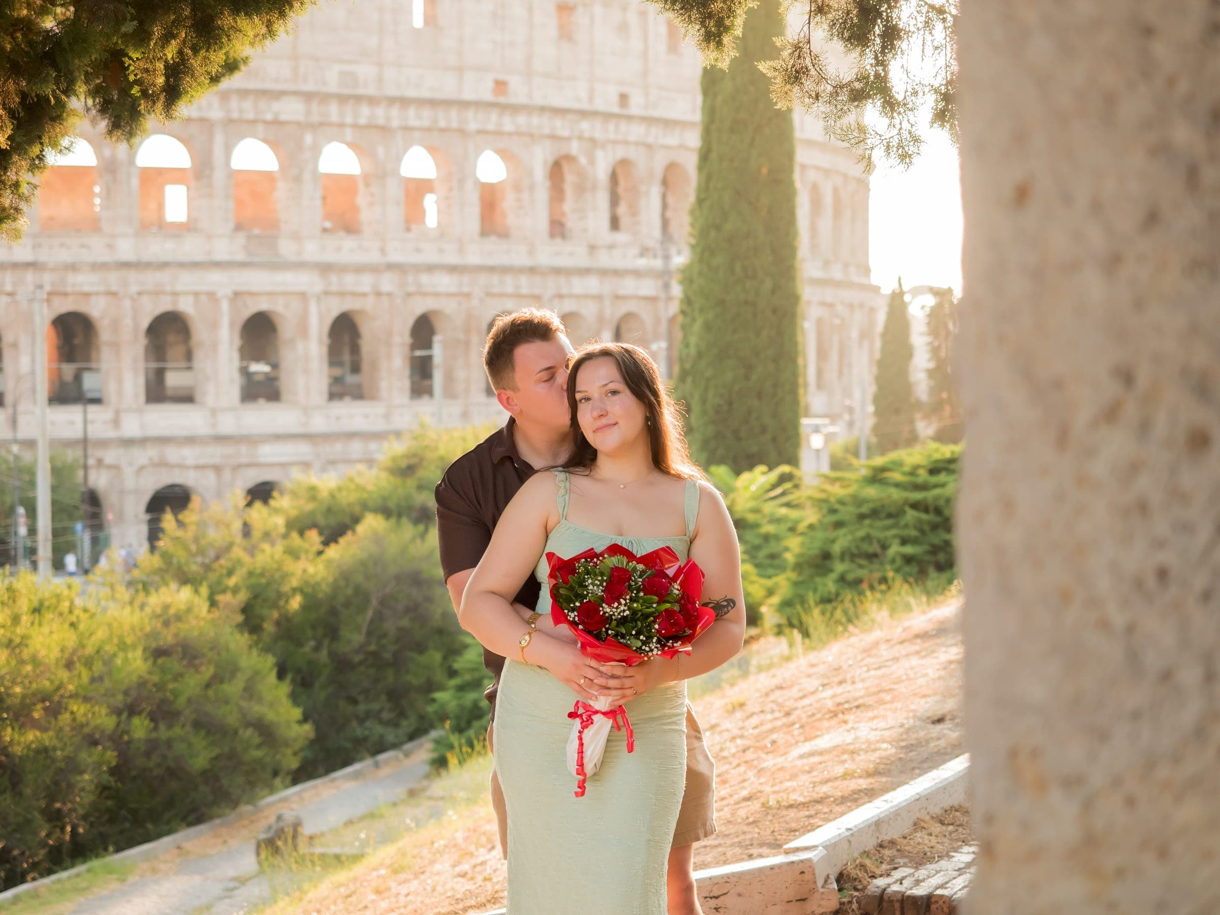 He kisses her temple as she holds red roses, the Colosseum in late sun behind them