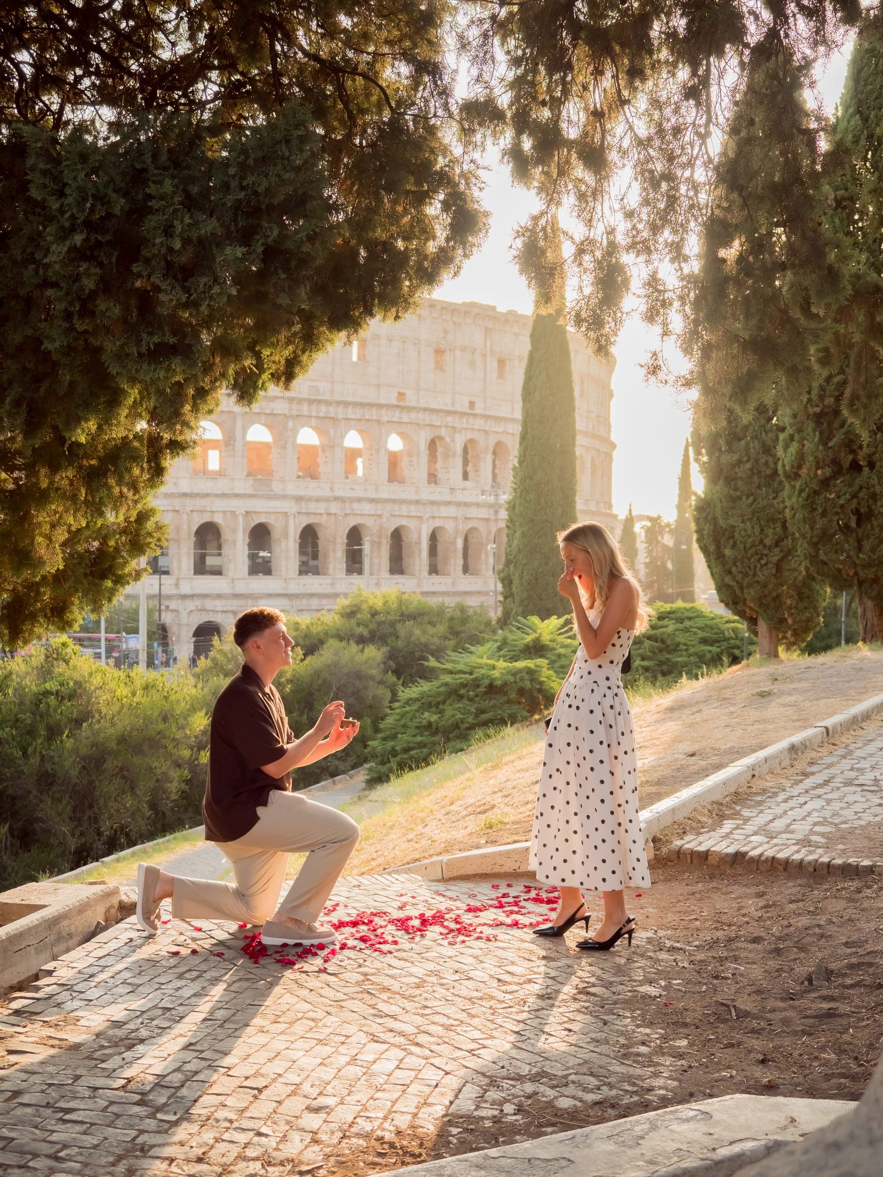 He kneels on rose petals under the pines, she covers her mouth, the Colosseum behind