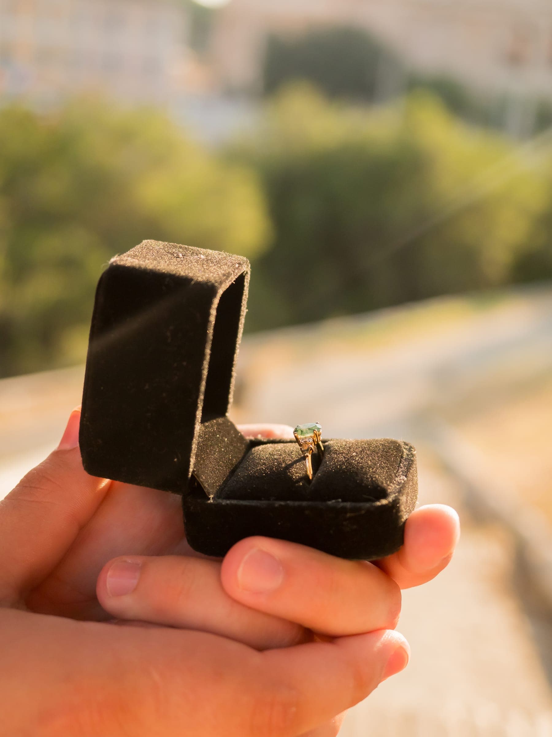 An open velvet ring box with an emerald engagement ring, held up in golden light